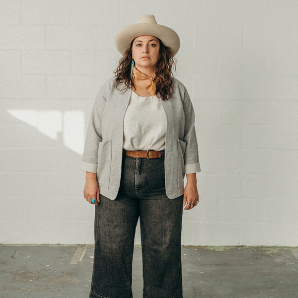 woman looking at camera standing in a white room wearing the denim artist jacket and a white top