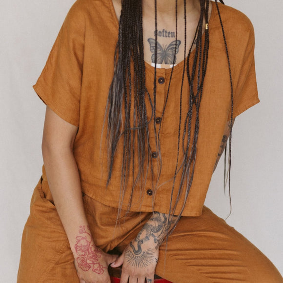 cropped image of woman wearing rust colored linen crop top with buttons on front and a butterfly chest tattoo and long braids