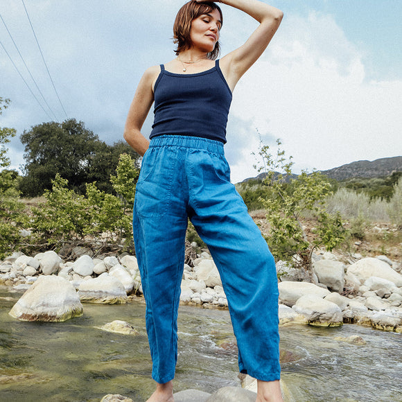 Woman in blue pants and navy top standing on a rock by a stream with trees and mountains in the background.