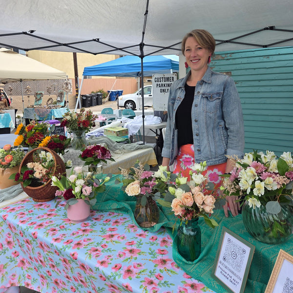 Woman standing behind a table with floral arrangements at an outdoor market.