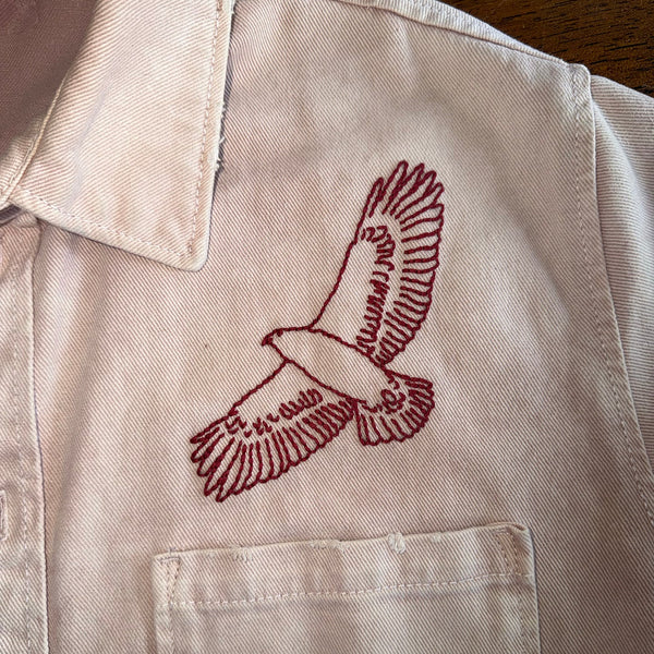 A close-up of a light beige or off-white shirt jacket with a red embroidered bird of prey design on the chest and a buttoned collar.