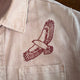 A close-up of a light beige or off-white shirt jacket with a red embroidered bird of prey design on the chest and a buttoned collar.