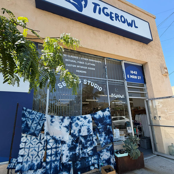 A storefront exterior for Tigerowl, with blue-and-white tie-dye textiles hanging outside the glass entrance. The building facade is beige, with a large white sign reading TIGEROWL and a blue logo.