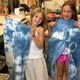 Two young girls smiling and holding up blue and white tie-dye garments in a sewing workshop. One girl wears a white tank top and beige polka-dot pants, and the other wears a white top, pink shorts, and black boots.