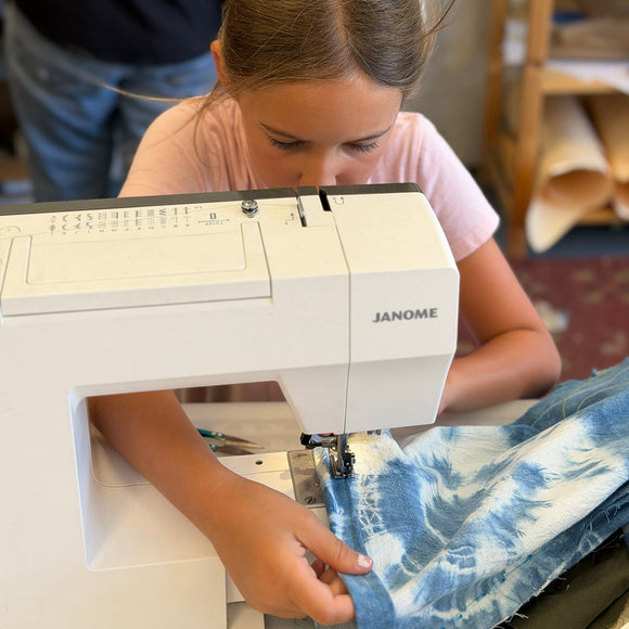 A young girl sewing a blue and white tie-dye fabric garment using a white Janome sewing machine in a workshop setting.