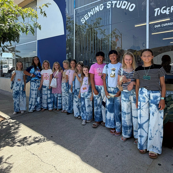 A group of young girls wearing matching blue and white tie-dye wide-leg pants stand together on a sidewalk in front of a storefront with large glass windows and the words 'SEwing STUDIO' and 'TIGER.