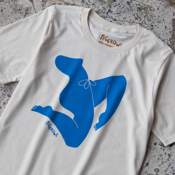 an image of a shirt laying on the ground. shirt is off white with a blue graphic of a lady's body in motion