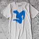 straight down shot of shirt on floor. shirt is off white with blue graphic of woman's figure in motion.