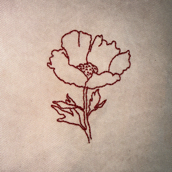 A close-up of a beige woven textile, likely a table runner or napkin, featuring a simple embroidered poppy flower in dark red thread.