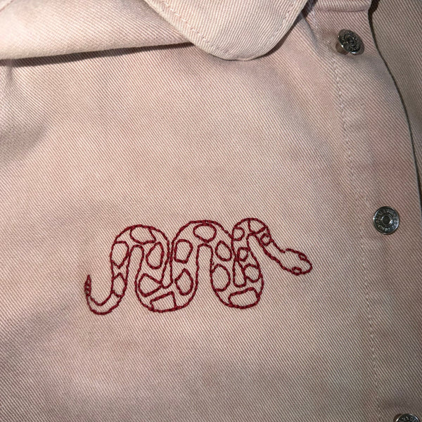 Close-up of a light beige or off-white collared shirt with a red embroidered snake motif on the chest and silver-toned buttons.