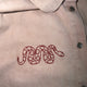 Close-up of a light beige or off-white collared shirt with a red embroidered snake motif on the chest and silver-toned buttons.