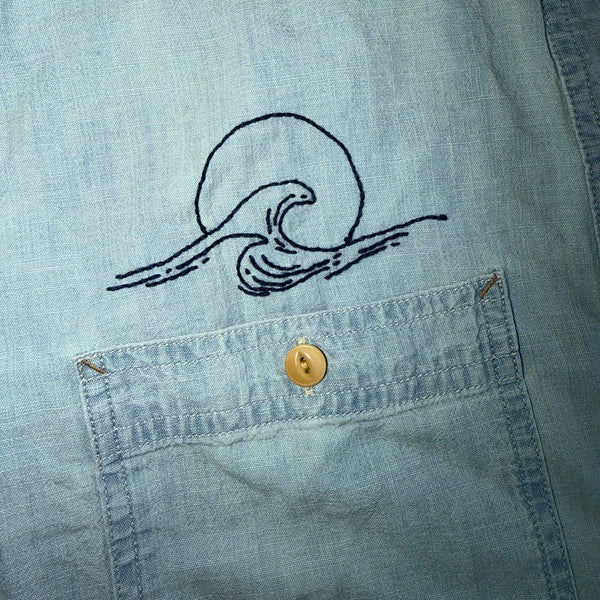 Close-up of a light blue denim shirt pocket with a black embroidered wave and sun design above a gold-tone button.