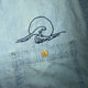 Close-up of a light blue denim shirt pocket with a black embroidered wave and sun design above a gold-tone button.