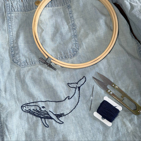 A light blue denim shirt laid flat with a whale embroidery design, shown with embroidery tools including a wooden hoop, scissors, a needle, and a spool of dark blue thread.
