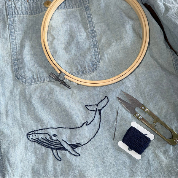 A light blue denim shirt laid flat with a whale embroidery design, shown with embroidery tools including a wooden hoop, scissors, a needle, and a spool of dark blue thread.