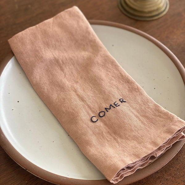 Beige napkin with 'comer' embroidery on a white plate on a wooden table