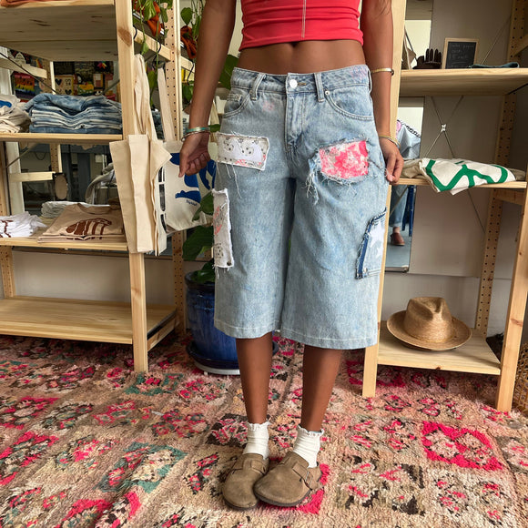 Person wearing a red top and patchwork denim shorts standing in a store with shelves and a patterned rug.