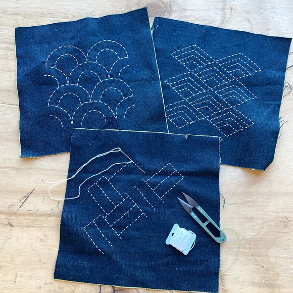 Three pieces of blue fabric with white embroidery on a wooden surface, accompanied by a pair of scissors.