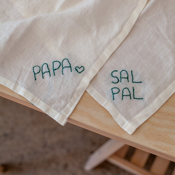 Two embroidered napkins on a wooden surface with 'PAPA' and 'SAL PAL' text.