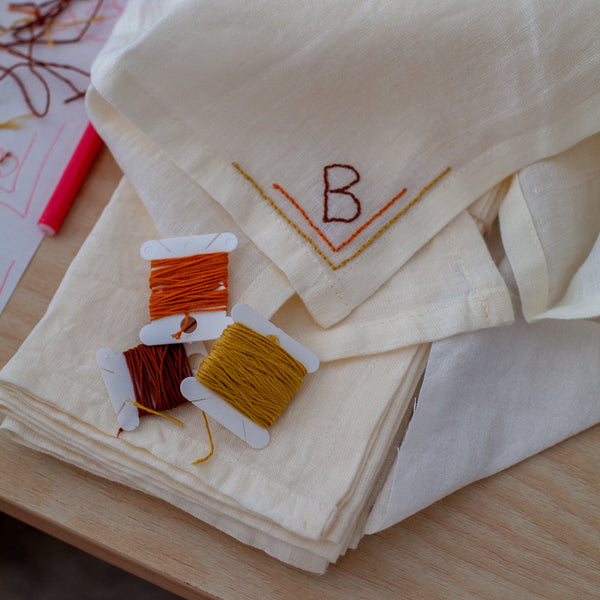 Stack of embroidered napkins with thread design and letter 'B' on a wooden surface