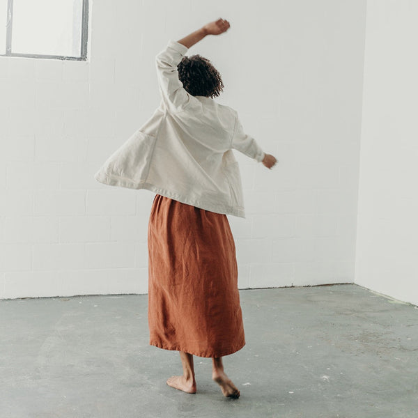 image of the back of a woman in a dancing position wearing cream colored artist jacket and rust colored linen skirt